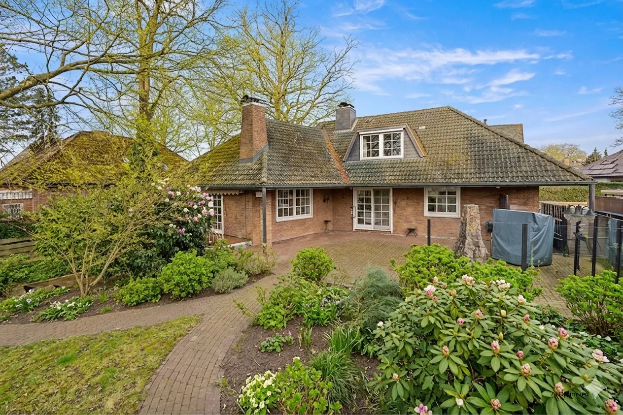 Brick house with a tiled roof, chimney, and white-framed windows. A brick patio and lush garden surround the house.