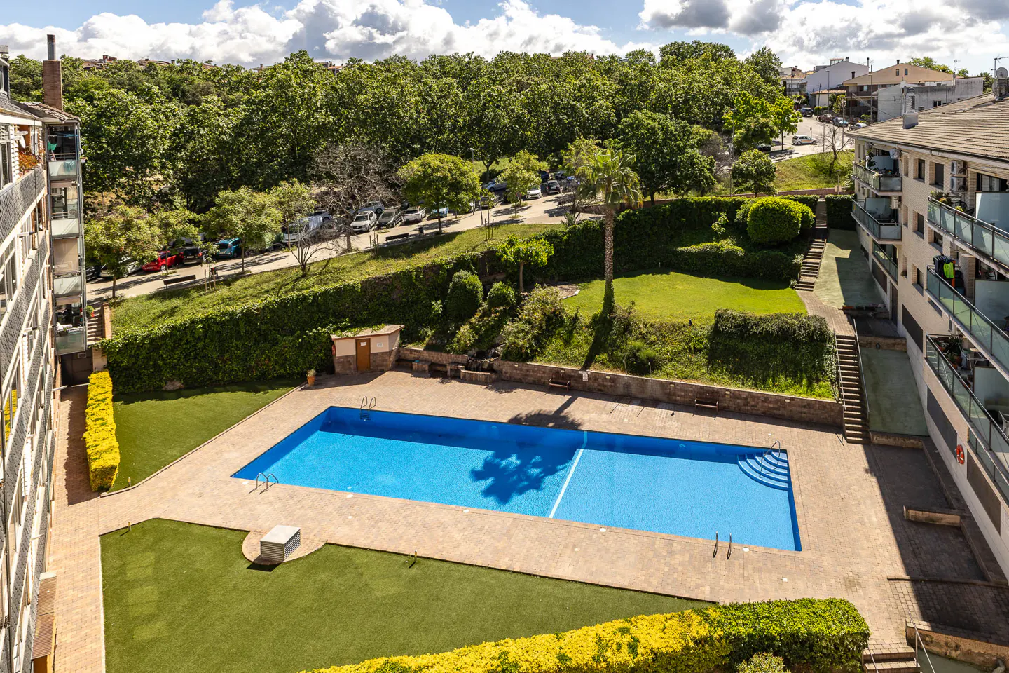 Aerial view of a blue swimming pool surrounded by green grass and trees, between two apartment buildings.