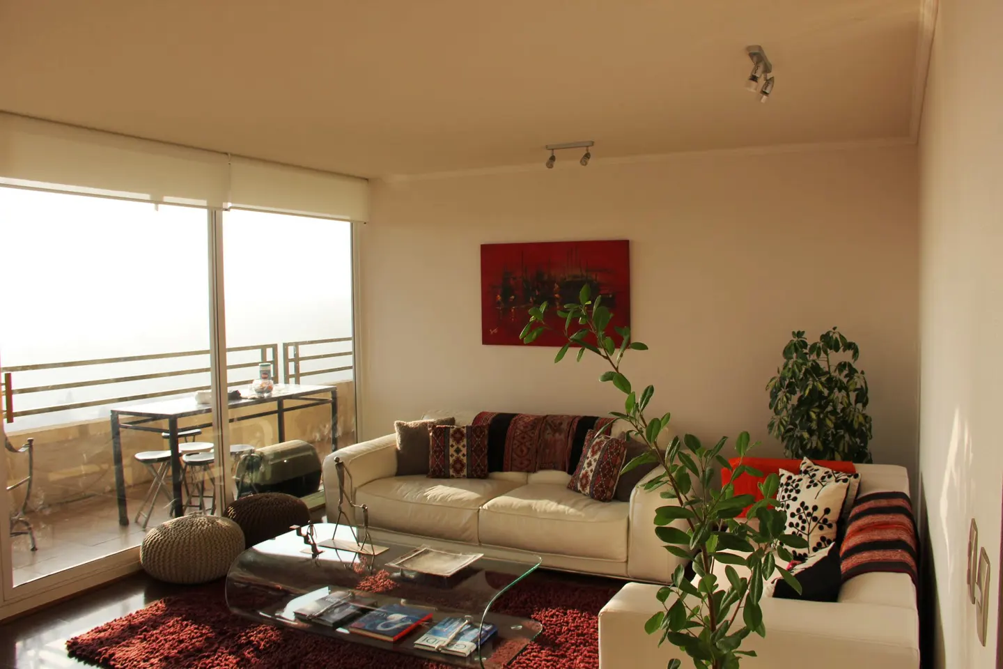 Bright living room with white sofas, patterned pillows, and a glass coffee table on a red rug. Balcony with table and chairs visible through sliding glass doors.