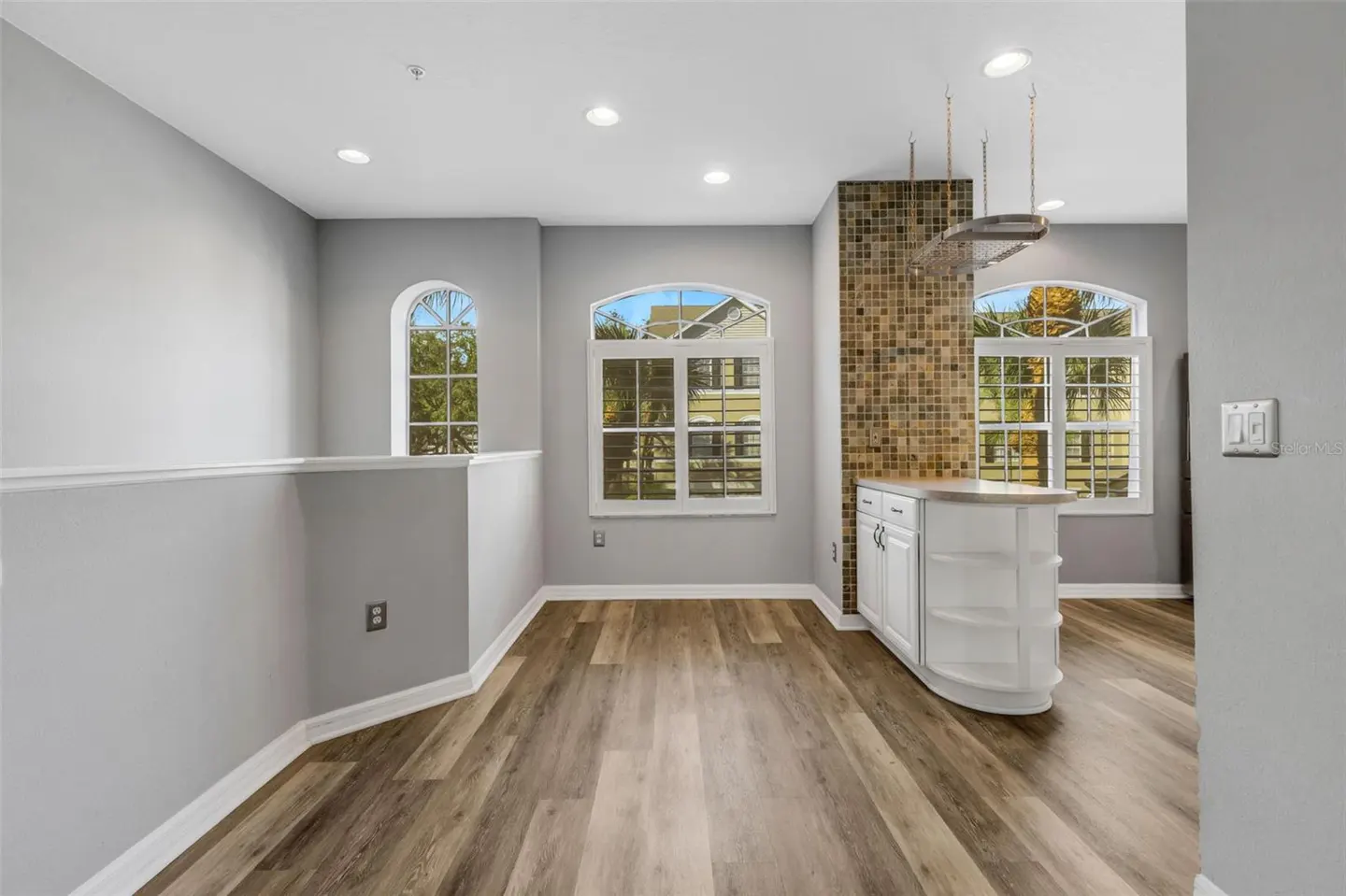 Bright, open-concept kitchen with wood floors, gray walls, and white cabinets. Three arched windows offer natural light. A mosaic tile backsplash adds texture.