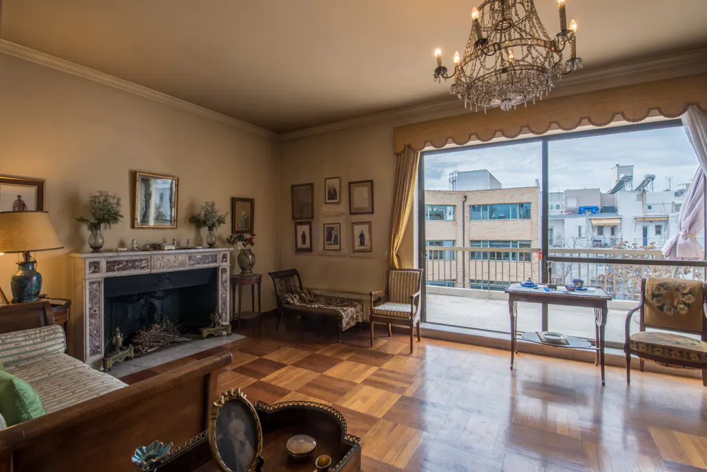 Elegant living room with parquet floors, a marble fireplace, and a crystal chandelier. A large window offers a city view.