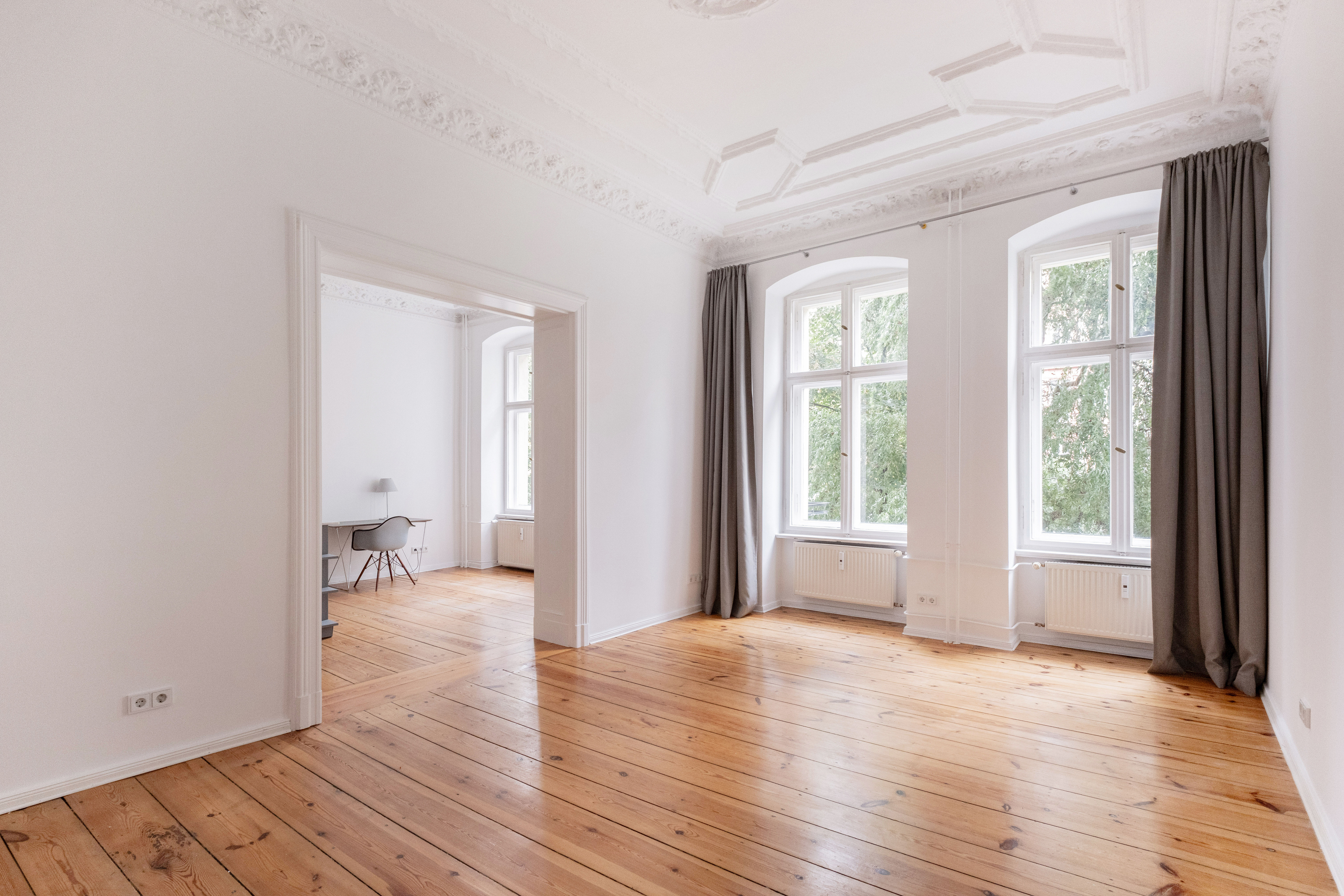 Bright, empty room with white walls, ornate ceiling molding, wood floors, and two large windows with grey curtains. [AI-generated text]
