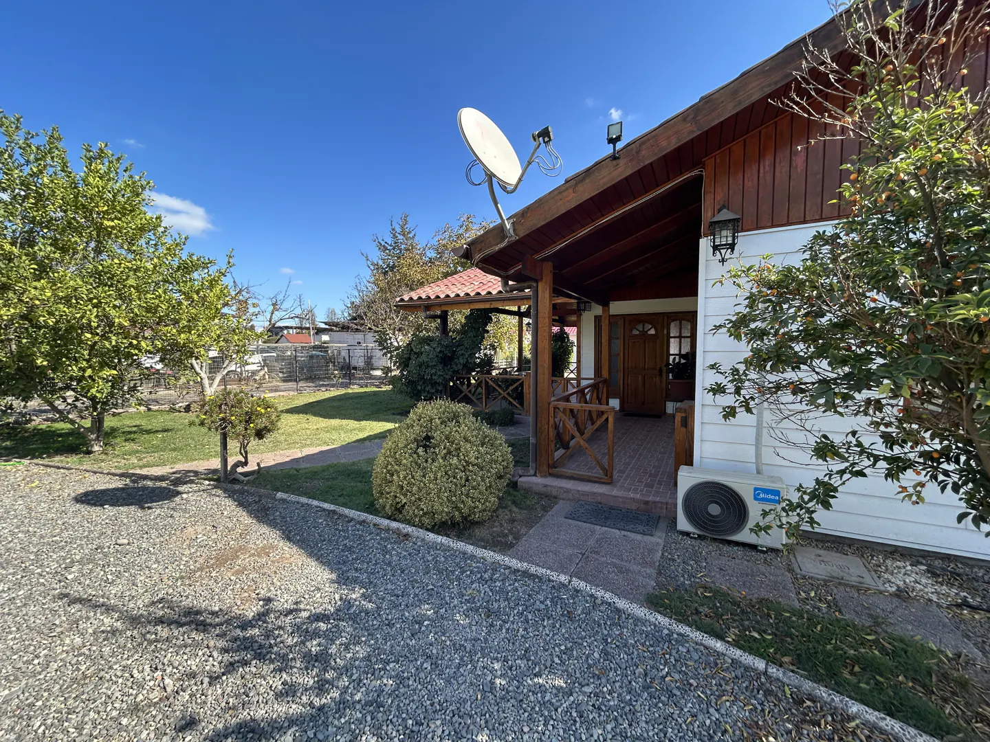 Exterior of a white house with a wood porch, satellite dish, and gravel driveway. Trees and blue sky in the background.