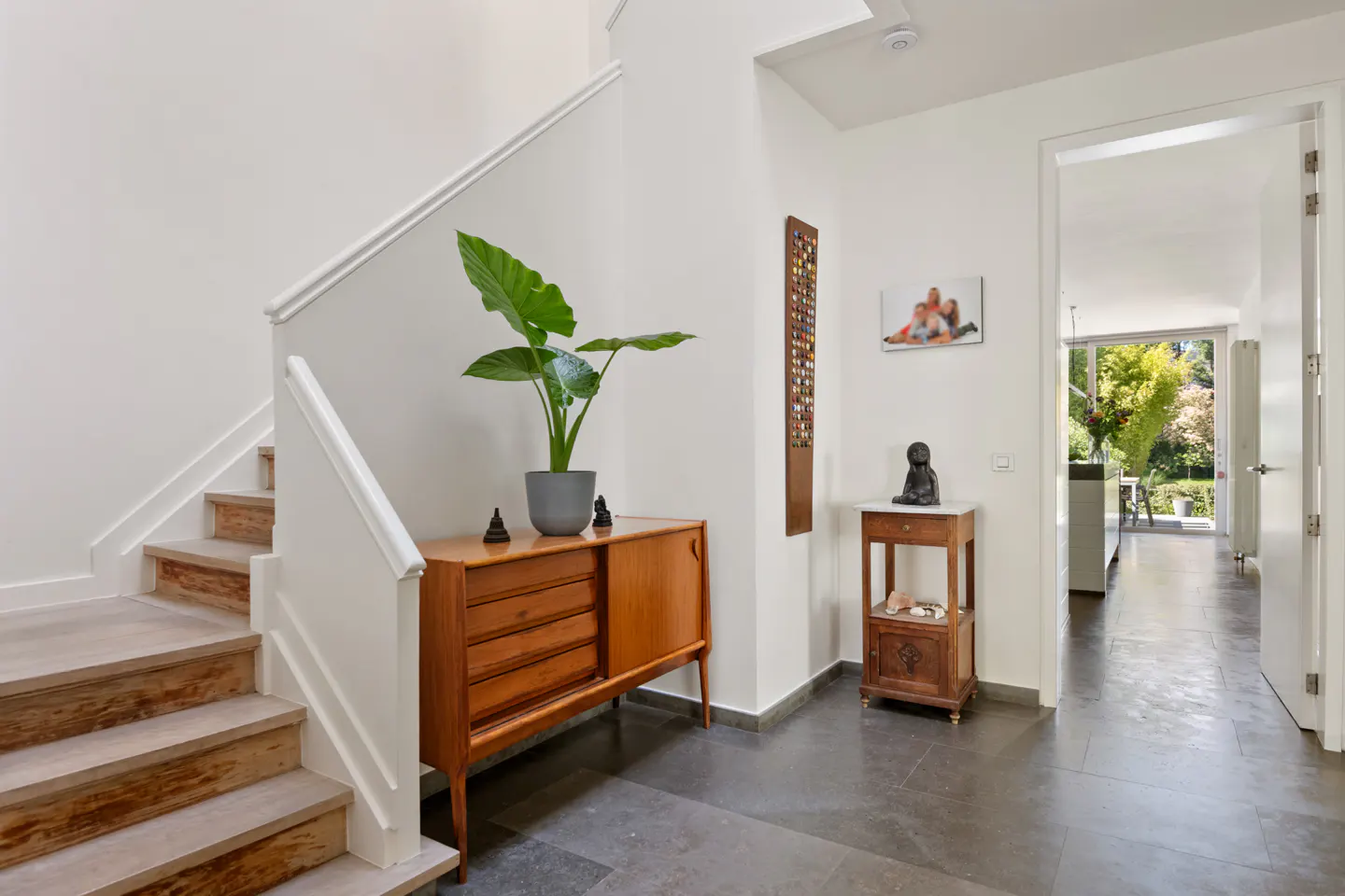 Bright entryway with stairs, wood cabinet with plant, and open doorway to a sunlit room.