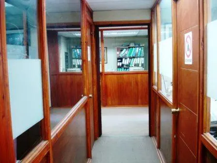 Hallway view of an office with wood-paneled walls and glass partitions. A doorway leads to another office with shelves of binders.