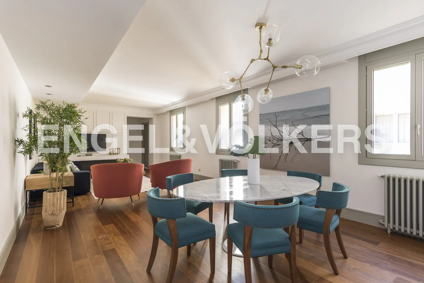 Bright, open-concept living and dining area with wood floors, teal chairs, and a marble table. A modern chandelier hangs above.