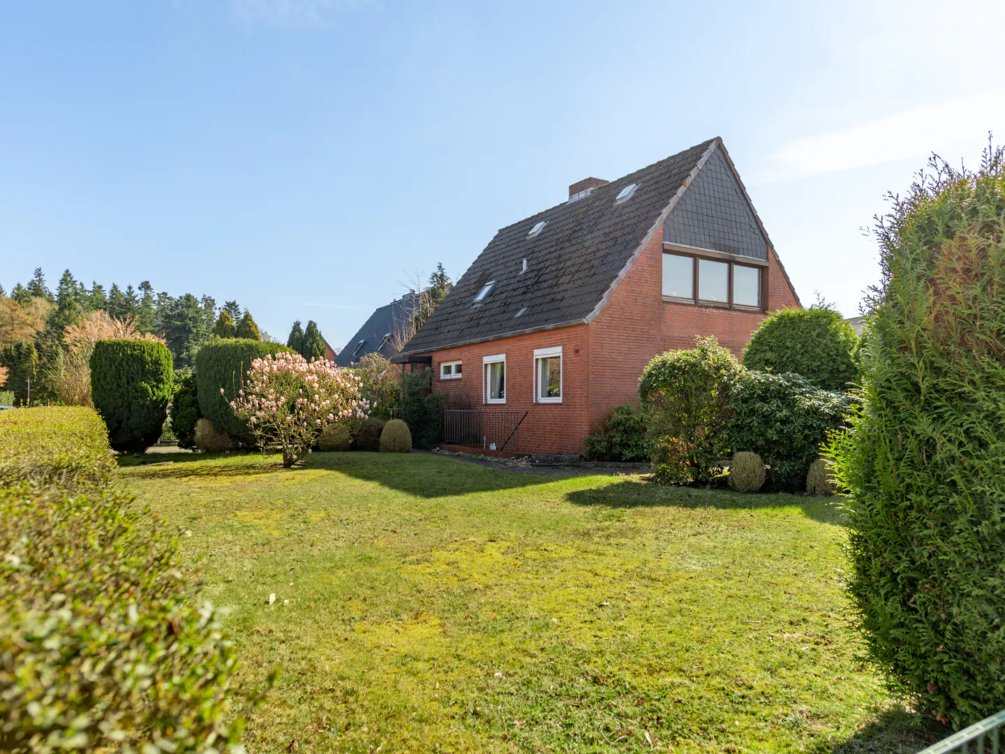 A red brick house with a gray roof sits on a green lawn with trees and bushes under a blue sky.