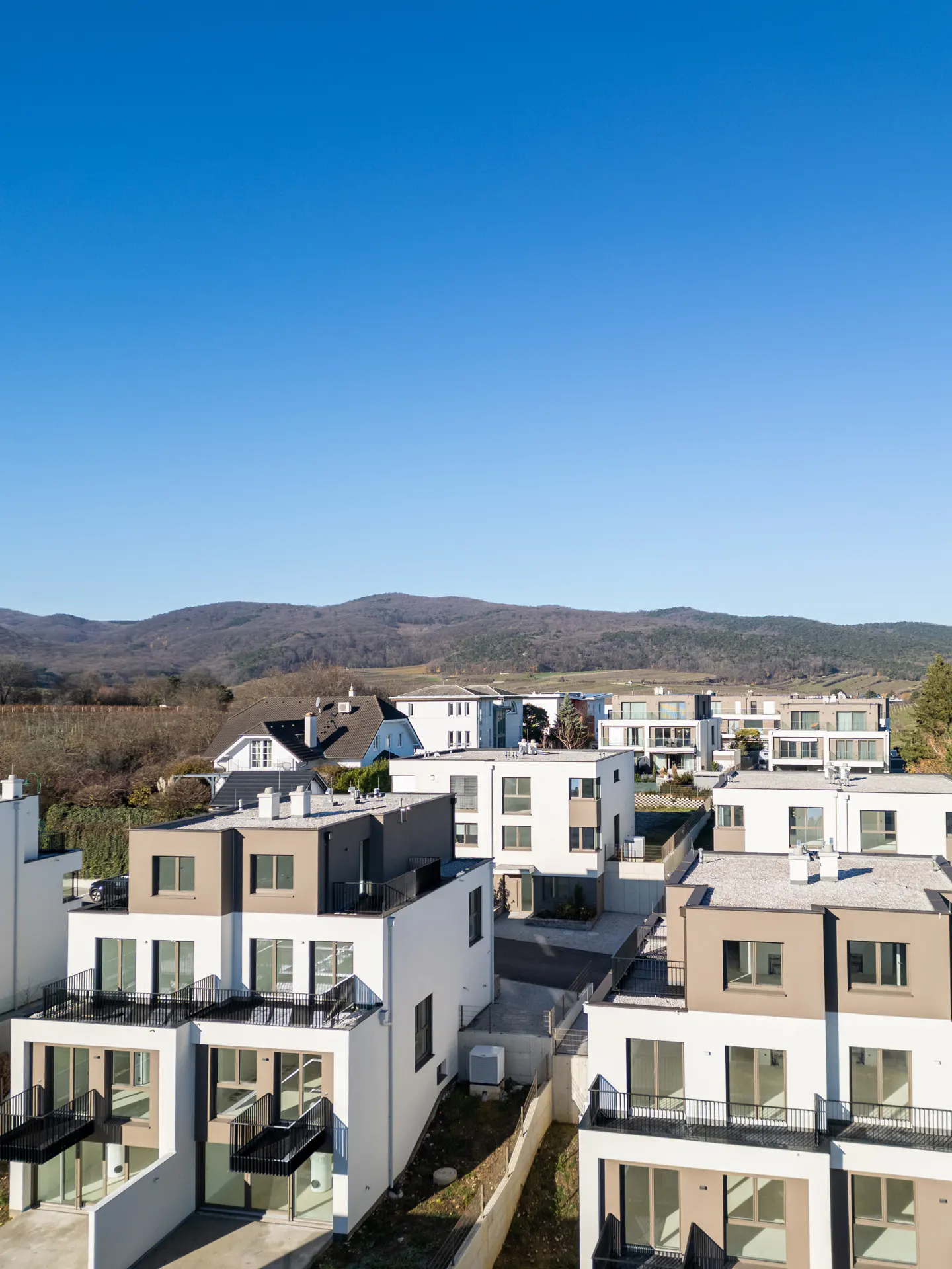 Aerial view of modern white and beige townhouses with black balconies under a clear blue sky, hills in the background.