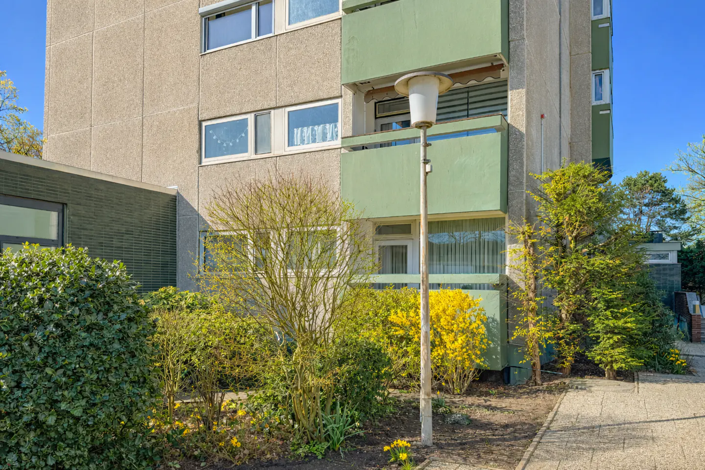 Exterior view of a multi-story apartment building with green balconies and a gray facade, surrounded by bushes and a lamp post.