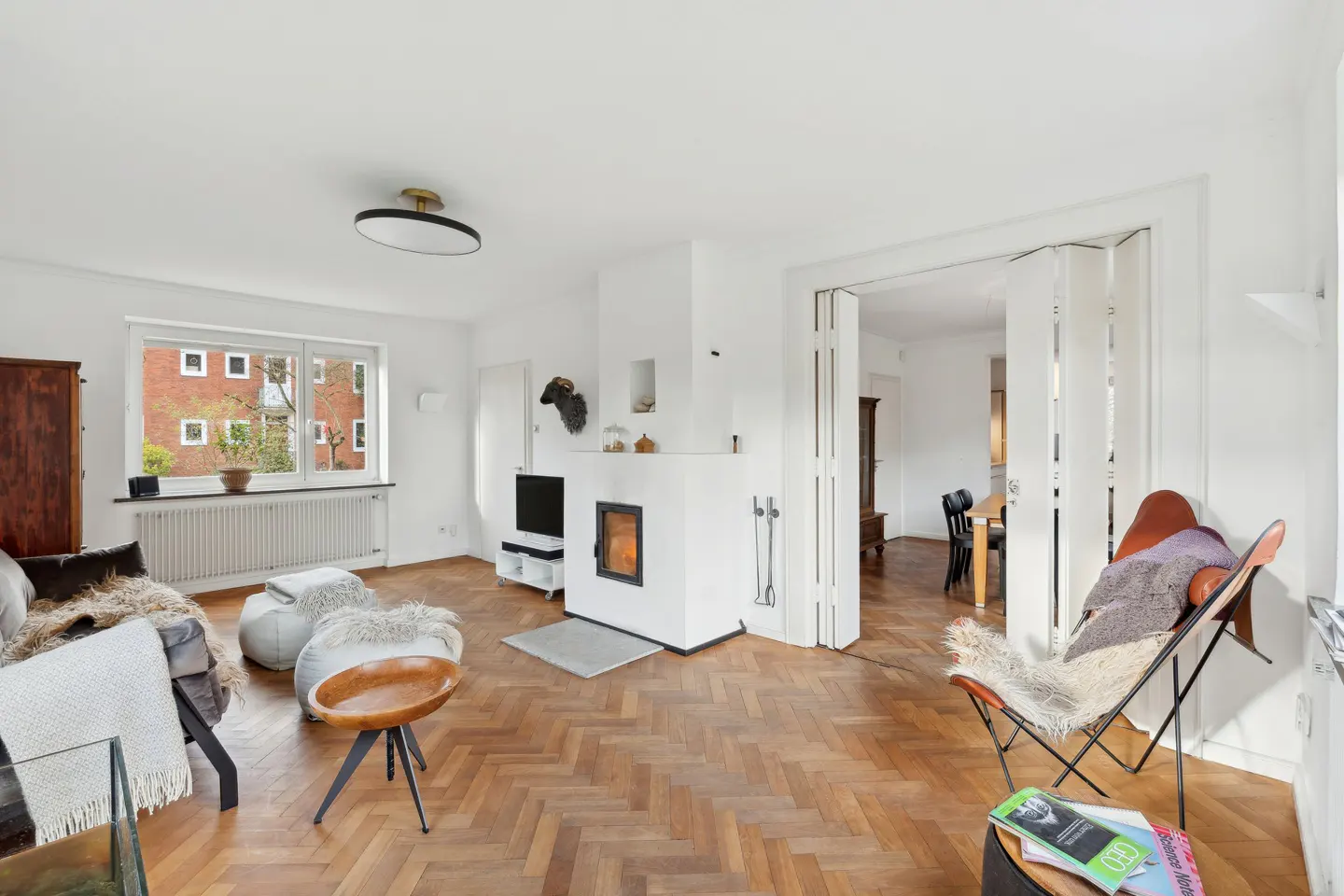 Bright living room with herringbone wood floors, white walls, and a fireplace. A window overlooks a brick building. Furniture includes a sofa, chairs, and a TV.