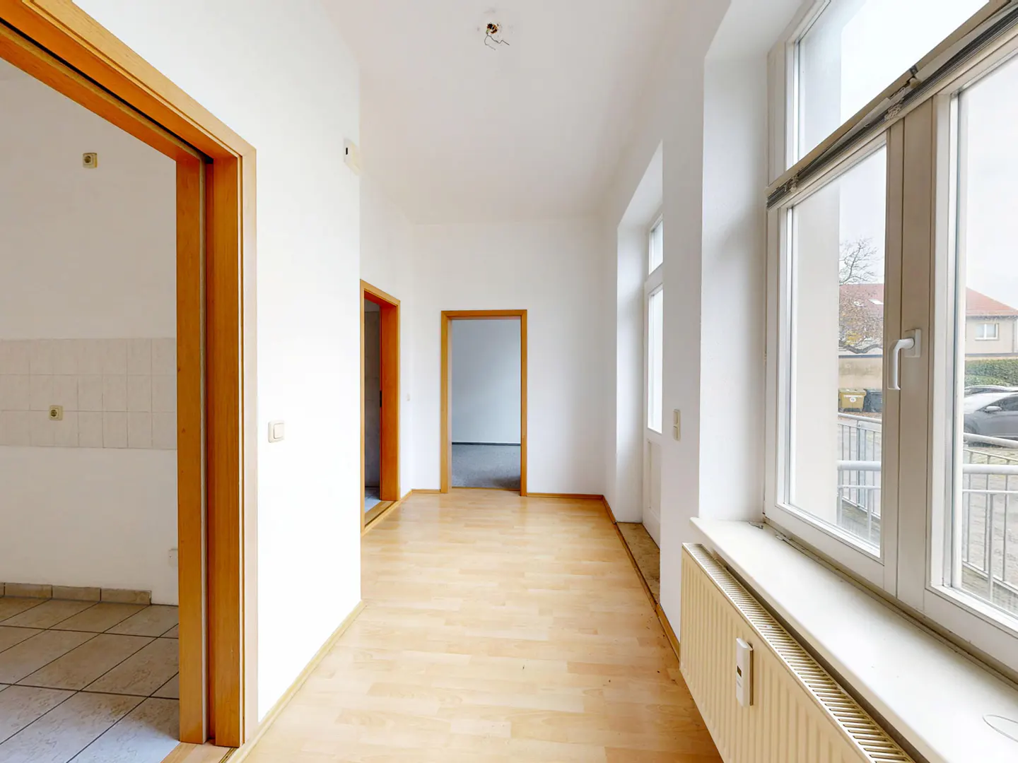Bright hallway with light wood floors, white walls, and wood-framed doorways. Large windows offer a view of the street.