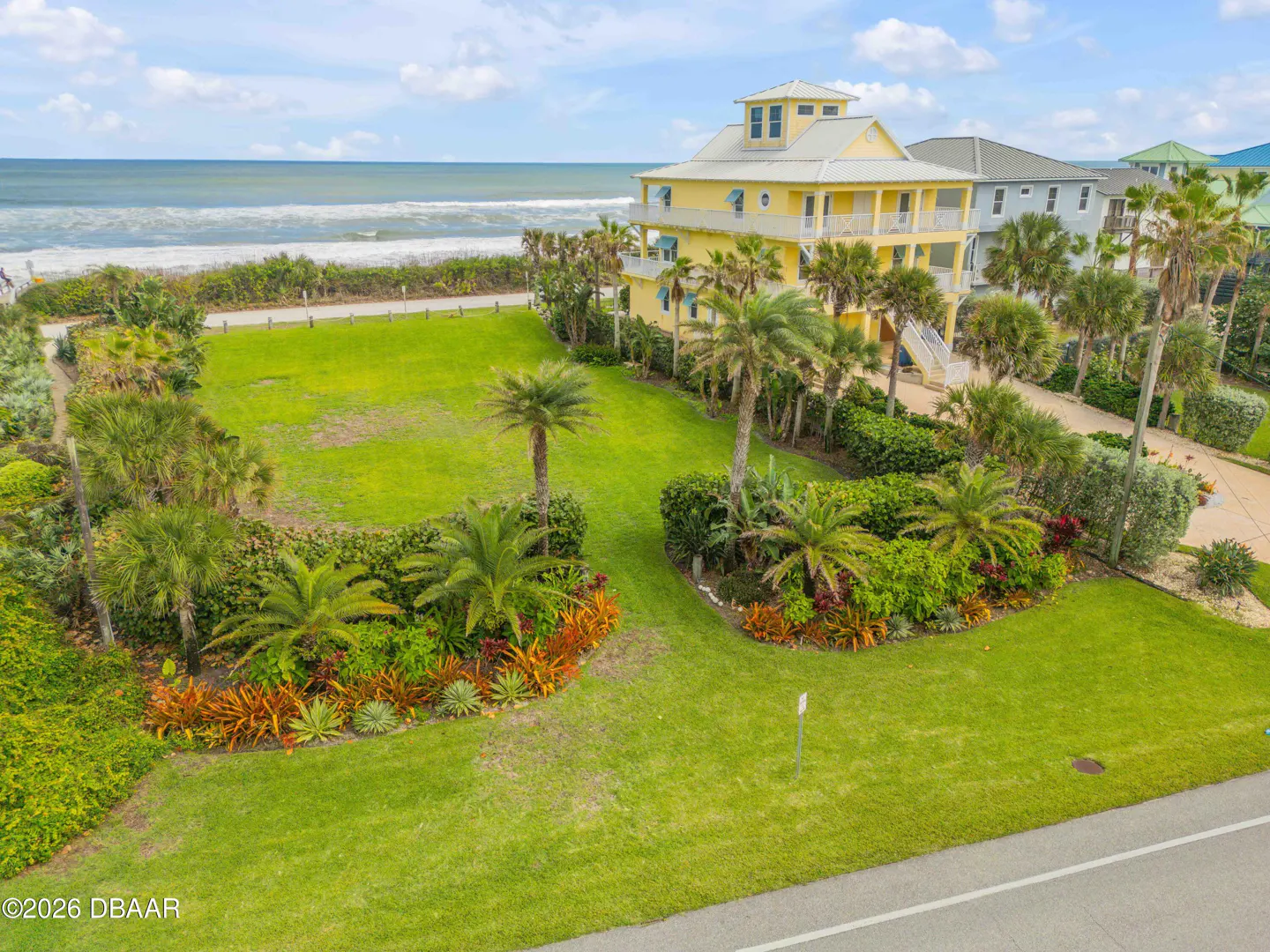 Aerial view of a yellow, multi-story beach house with palm trees and a green lawn, ocean in the background.