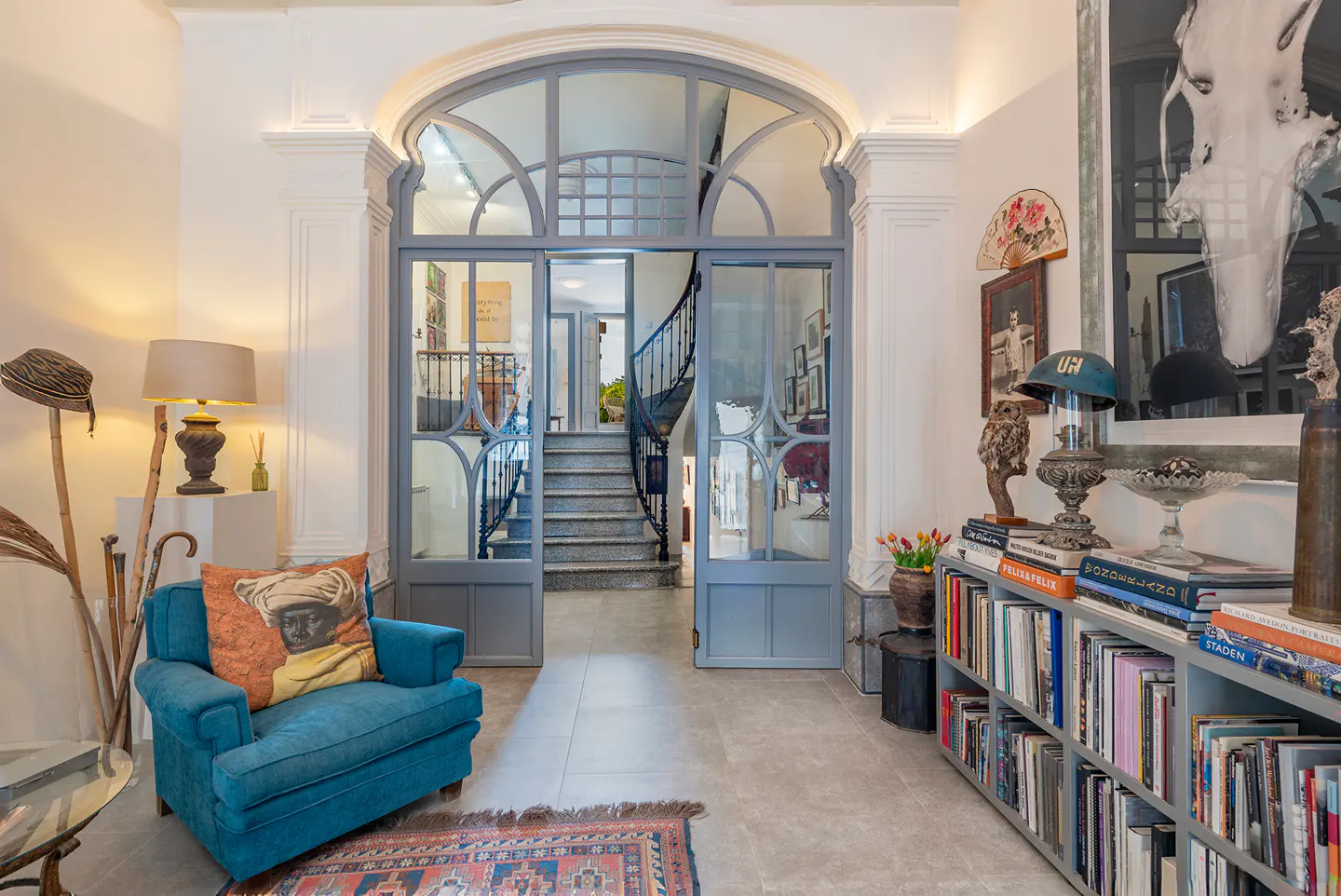 Elegant foyer with blue armchair, patterned pillow, and arched doorway leading to a staircase. Bookshelves and decor add character.