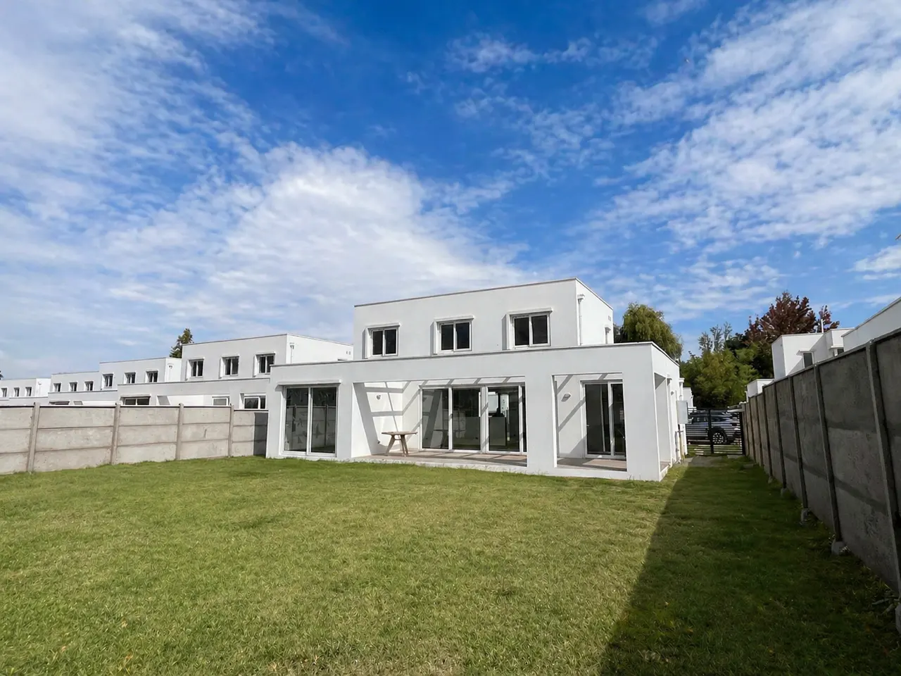 Exterior view of modern white townhouses with green lawn and concrete fence under a blue sky with clouds.