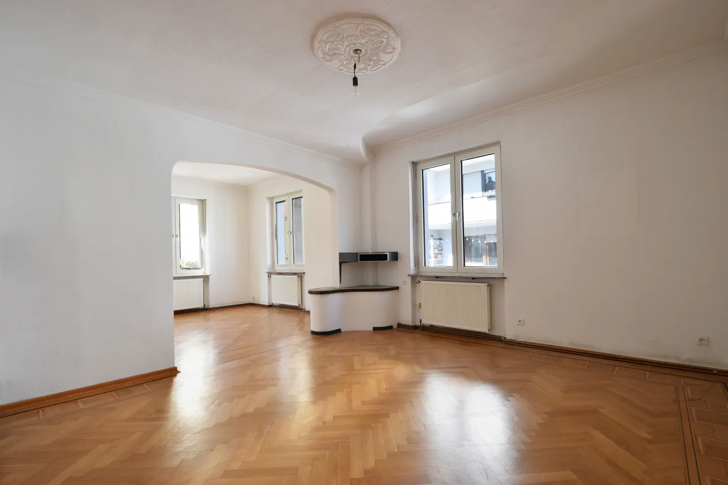 Bright, empty room with herringbone wood floors, white walls, and two windows. An archway leads to another room with a window.
