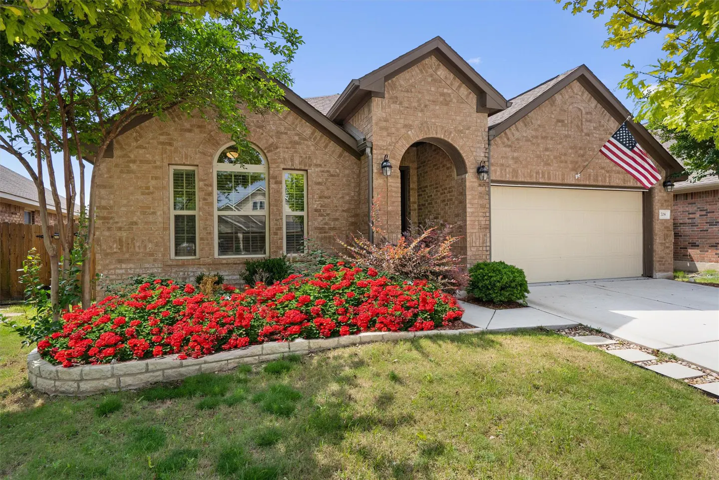 Tan brick house with an American flag, a beige garage door, and a flower bed with red flowers.