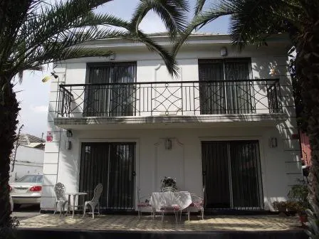 Two-story white house with a black iron balcony. A table and chairs sit on the patio, framed by palm trees.