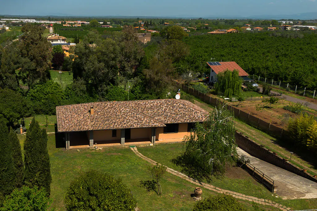 Aerial view of a single-story, peach-colored house with a terracotta tile roof, surrounded by green lawn and trees. A path leads to the house.