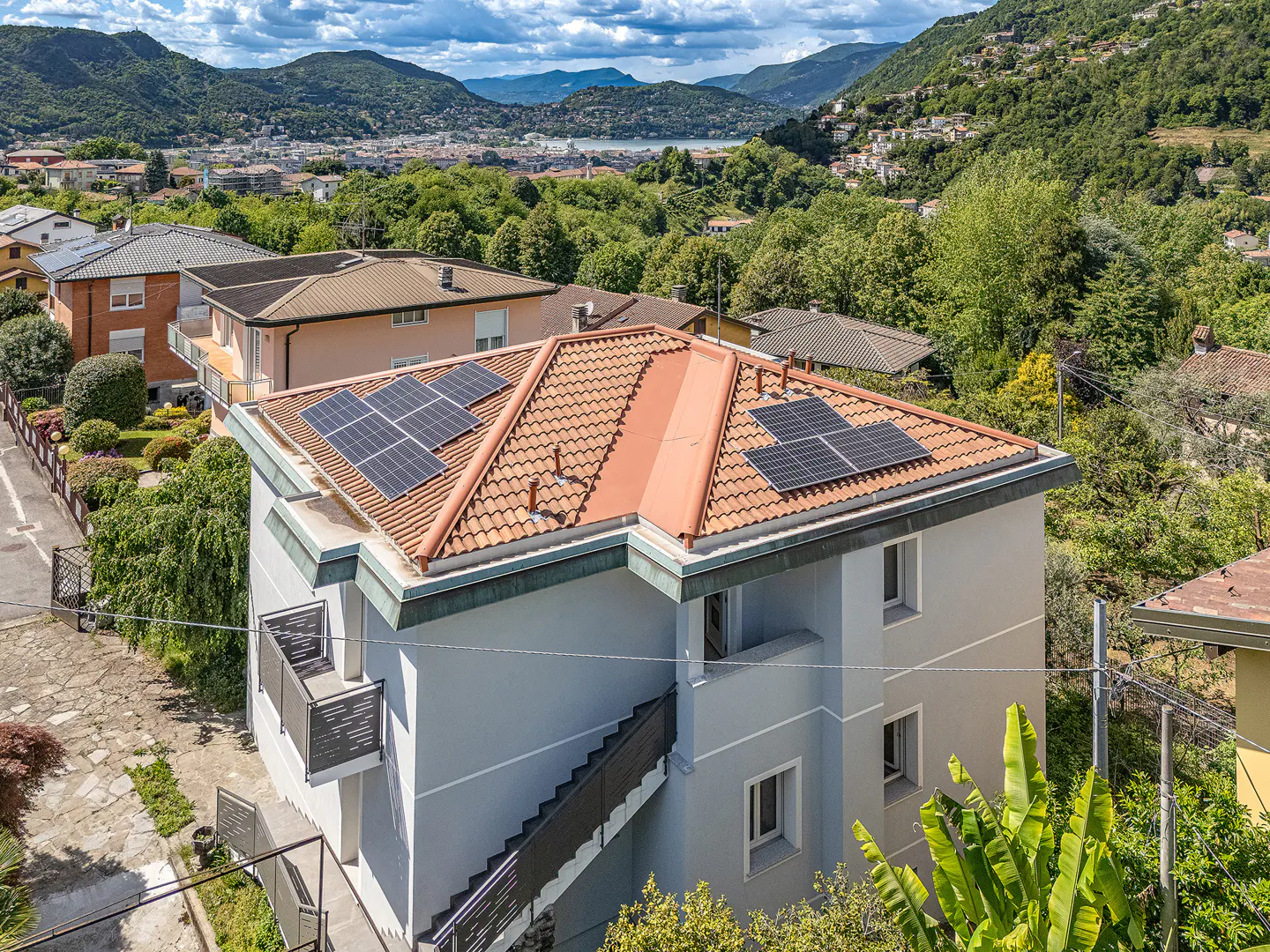 Aerial view of a gray house with solar panels on a red tile roof, set against a backdrop of green hills and a blue sky.