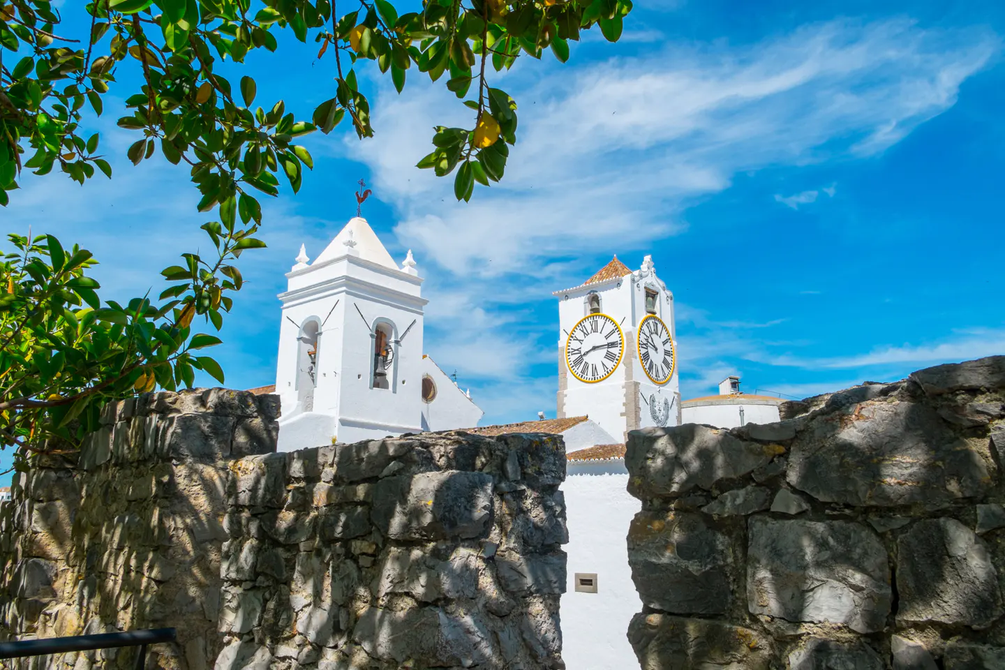 View of the Igreja Matriz clock tower in Tavira, Portugal, framed by a stone wall and green foliage against a blue sky.