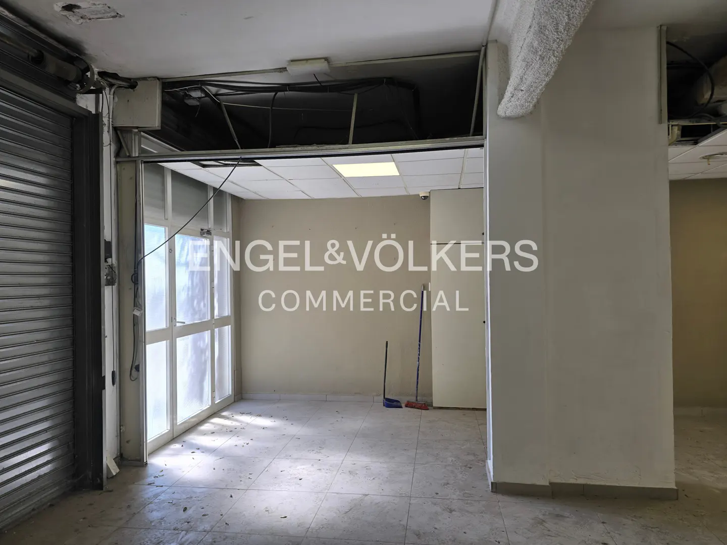Empty commercial space with tile floors, white walls, and Engel & Völkers Commercial logo. A metal shutter and glass doors are on the left.