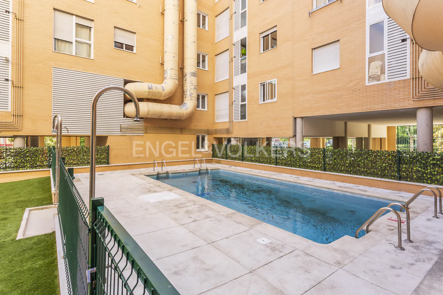 Outdoor swimming pool with blue water, surrounded by a stone patio, green privacy fencing, and tan brick apartment walls.