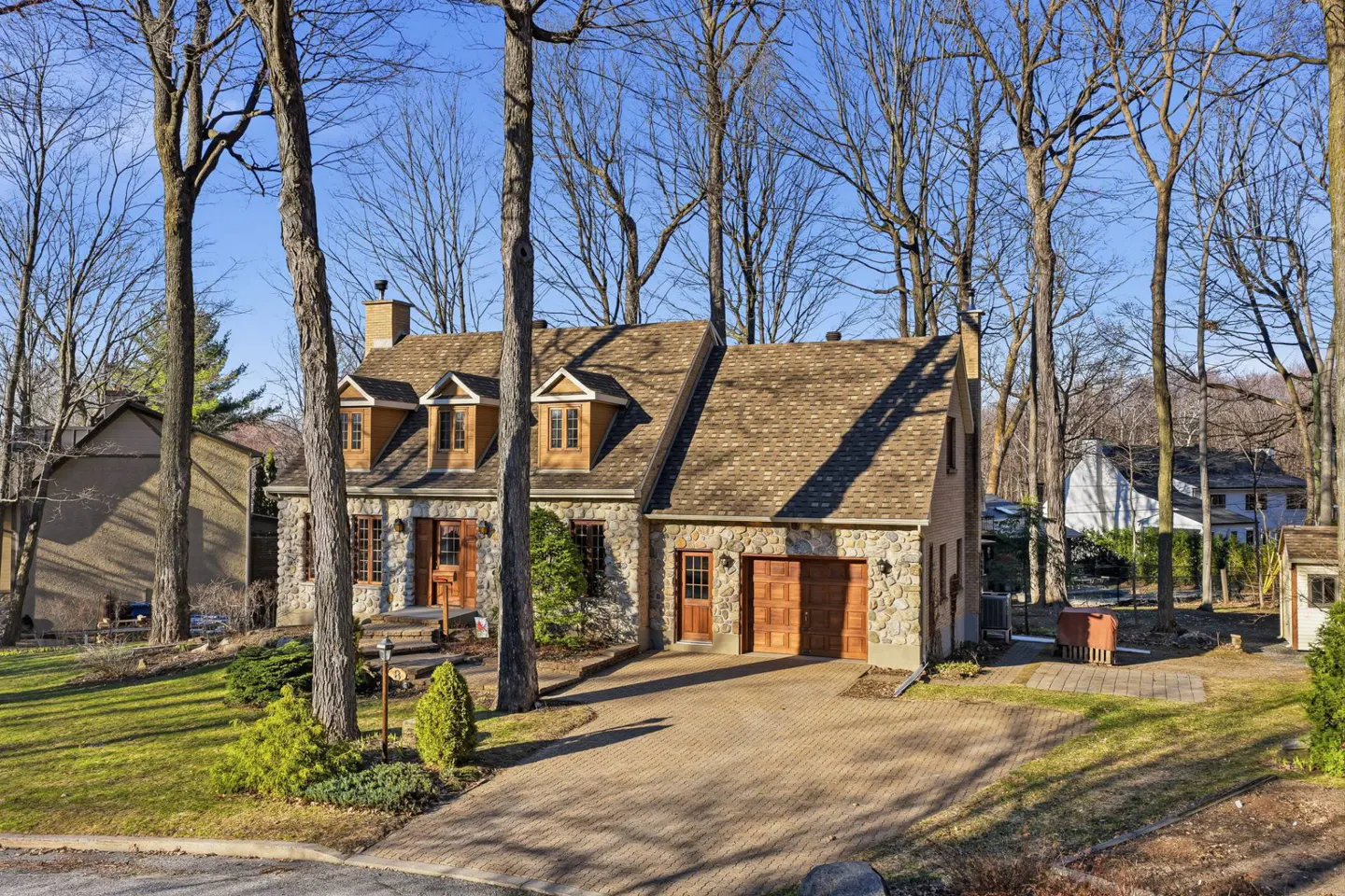 Stone house with brown roof and dormers, surrounded by trees. A brick driveway leads to a wooden garage door.