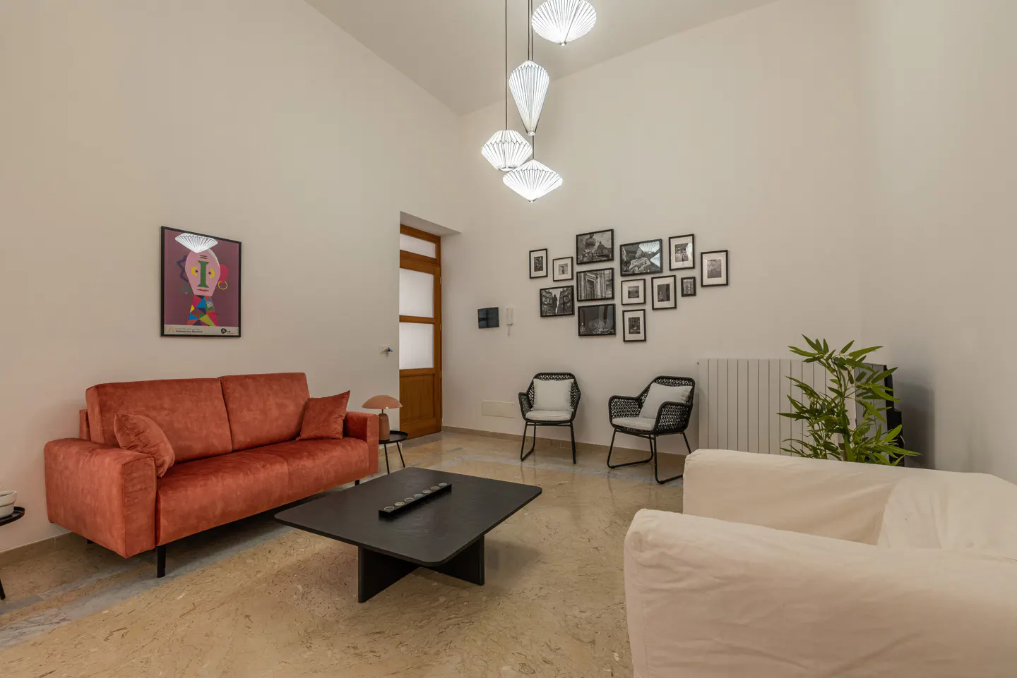 Bright living room with orange sofa, black coffee table, and white chairs. Black and white photos hang on the wall. Three pendant lights illuminate the space.