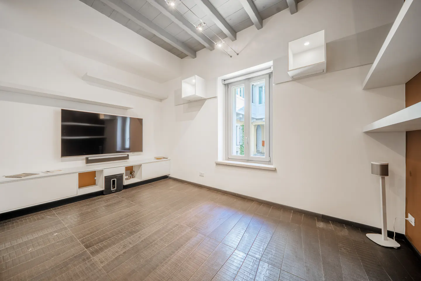 A modern living room with white walls, a large TV, and dark wood floors. A window lets in natural light.
