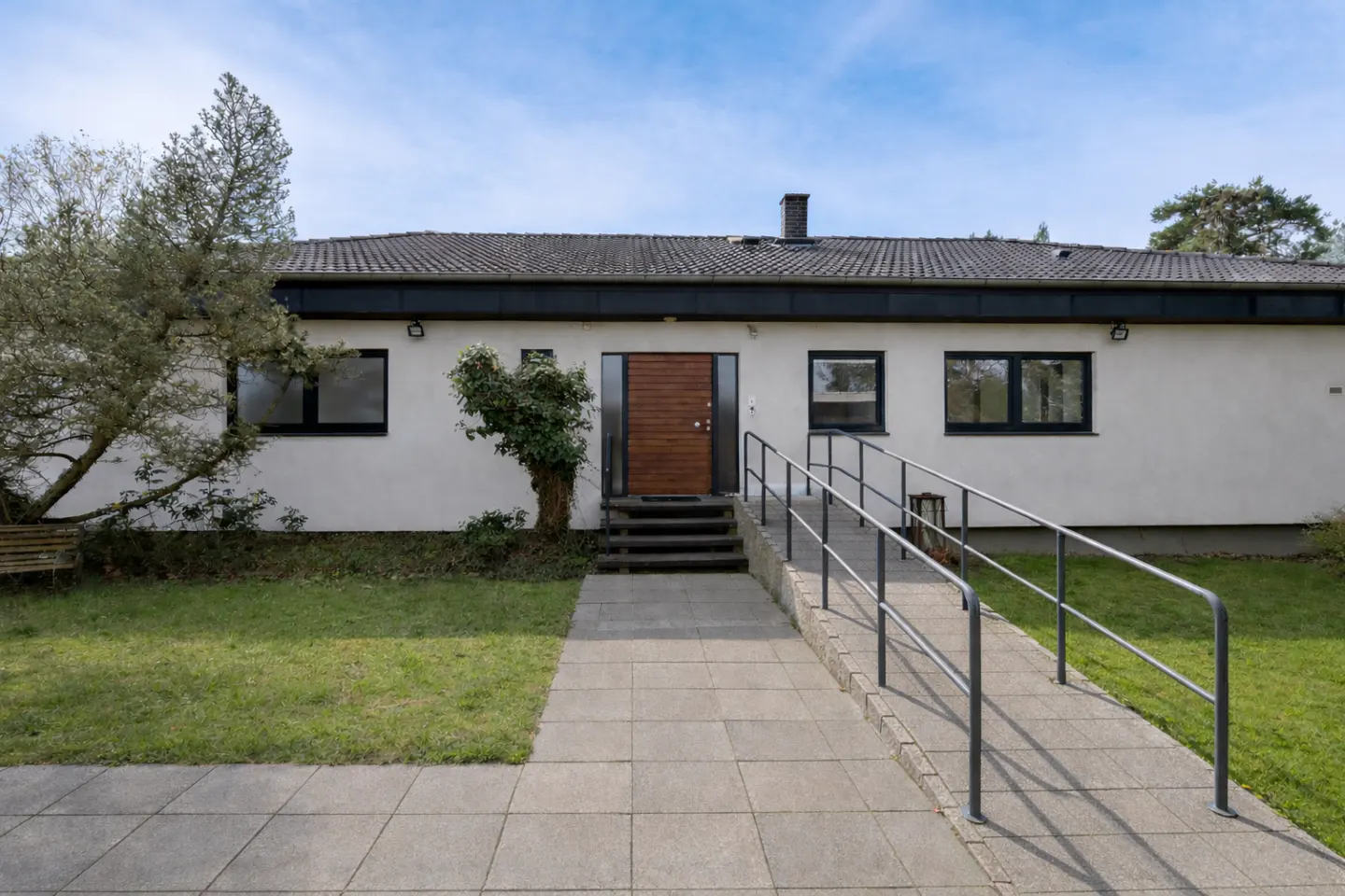 Exterior view of a one-story white house with a brown door, black trim, and a ramp with railings leading to the entrance.