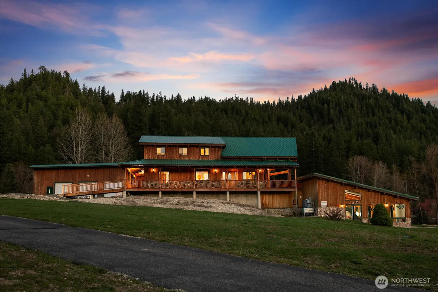 A large, two-story wooden house with a green roof sits on a grassy hill against a backdrop of forested mountains at dusk.