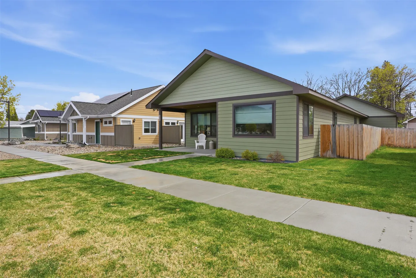 A green, single-story house with a brown roof and trim sits on a green lawn next to a sidewalk. Other houses are visible in the background.