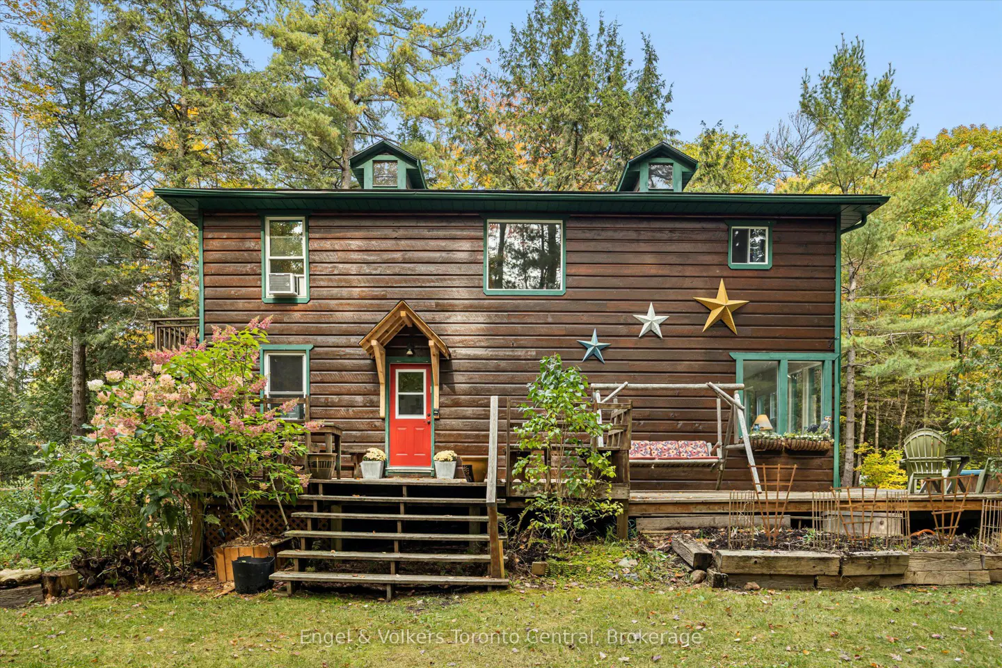 Two-story log cabin with a red door, green trim, and star decorations. A wooden deck and stairs lead to the entrance. Trees surround the house.