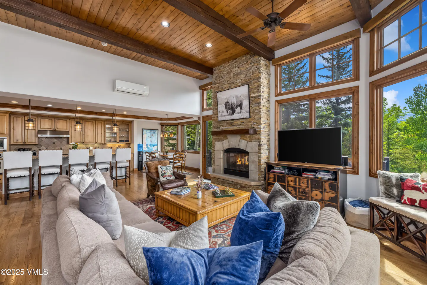 Living room with stone fireplace, wood ceiling, and large windows showing trees. A large sofa with blue pillows is in the foreground.