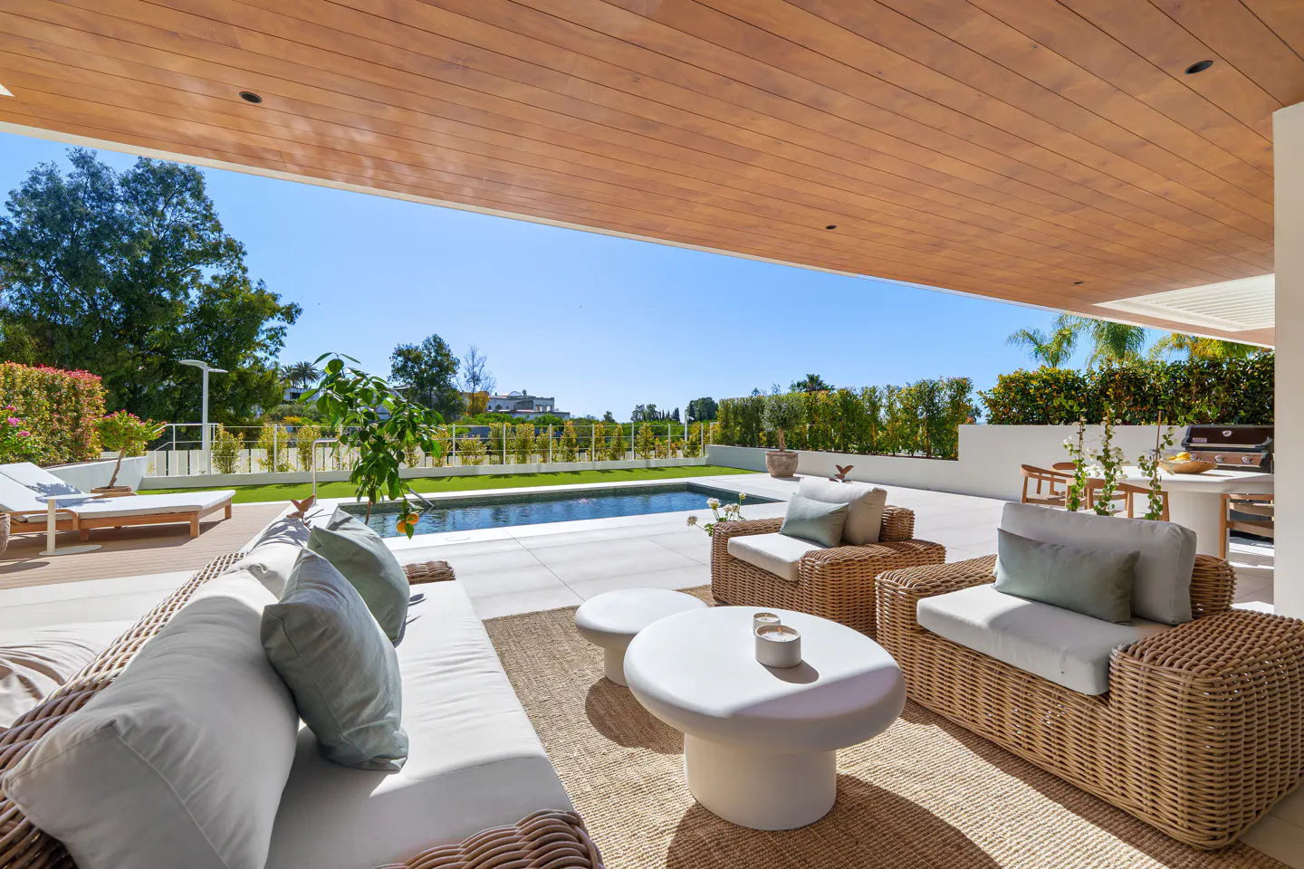 Outdoor patio with wicker furniture, white cushions, and a white table. A pool and green trees are in the background.
