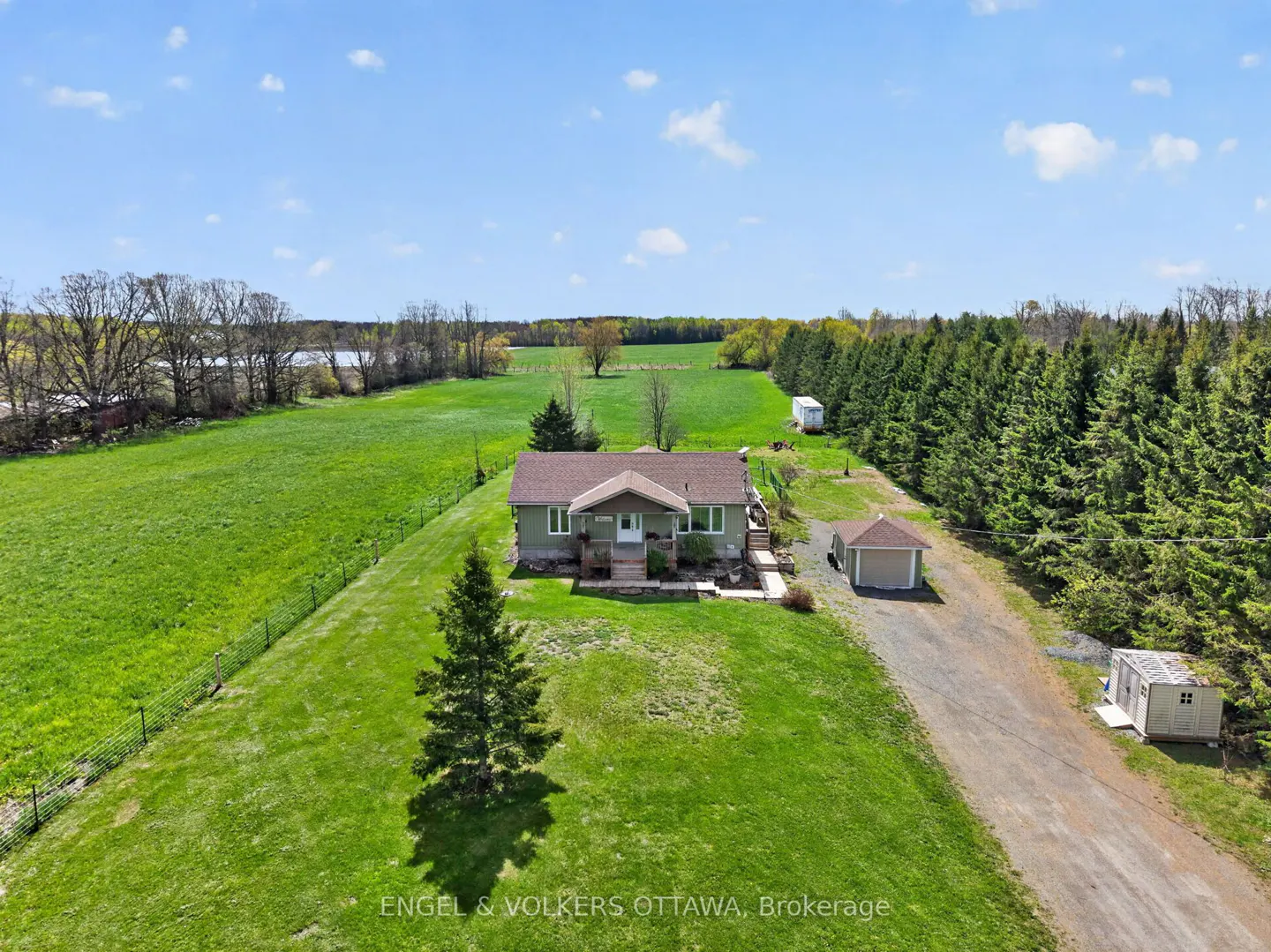 Aerial view of a sage green house with a brown roof, surrounded by green fields and trees under a blue sky.