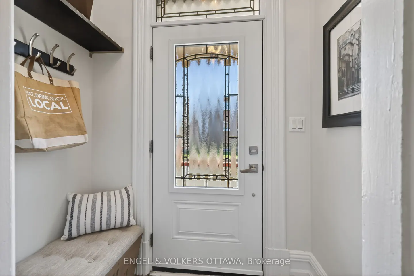 Entryway with a white door with decorative glass, a bench with a striped pillow, and a tote bag hanging on hooks.