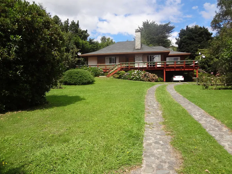 Exterior view of a brown house with a stone chimney, red deck, and stone driveway on a green lawn. A white car is parked under the deck.