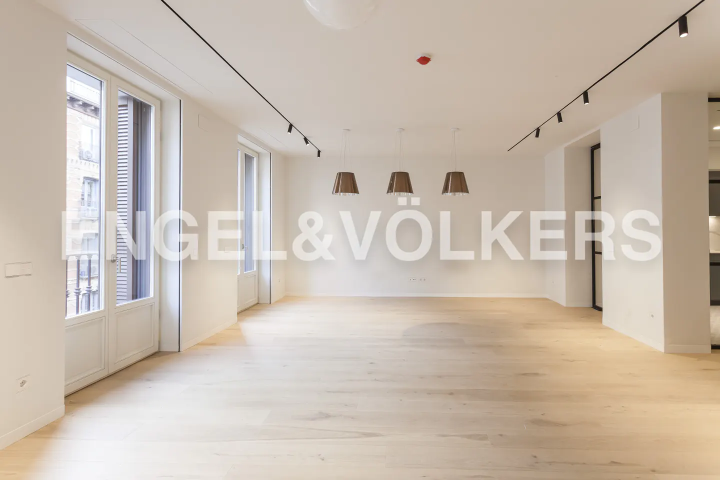 Bright, empty room with light wood floors, white walls, and three pendant lights. Windows and track lighting add to the modern feel.