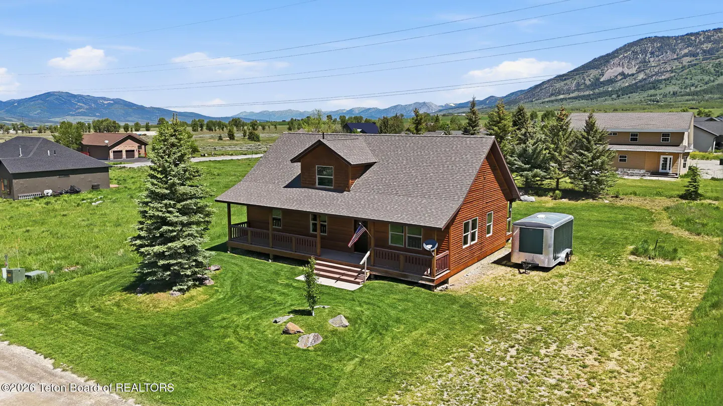 Aerial view of a brown log cabin with a porch and American flag, set in a grassy field with mountains in the background.