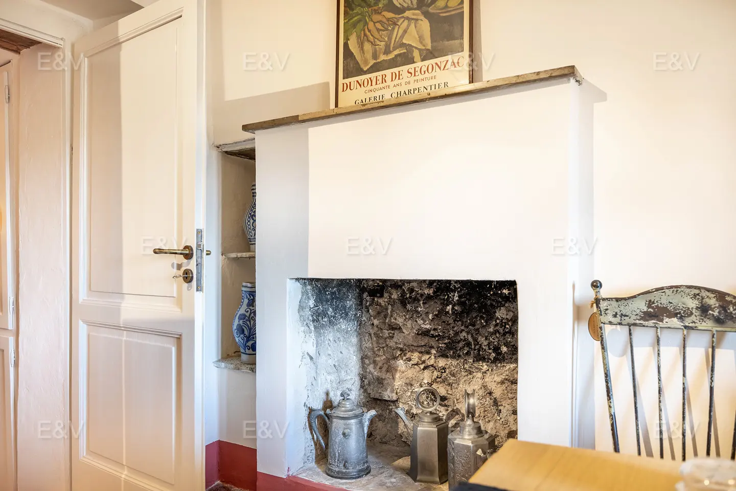 Interior view of a white fireplace with metal pots, a vintage chair, and a white door with a shelf holding blue and white vases.