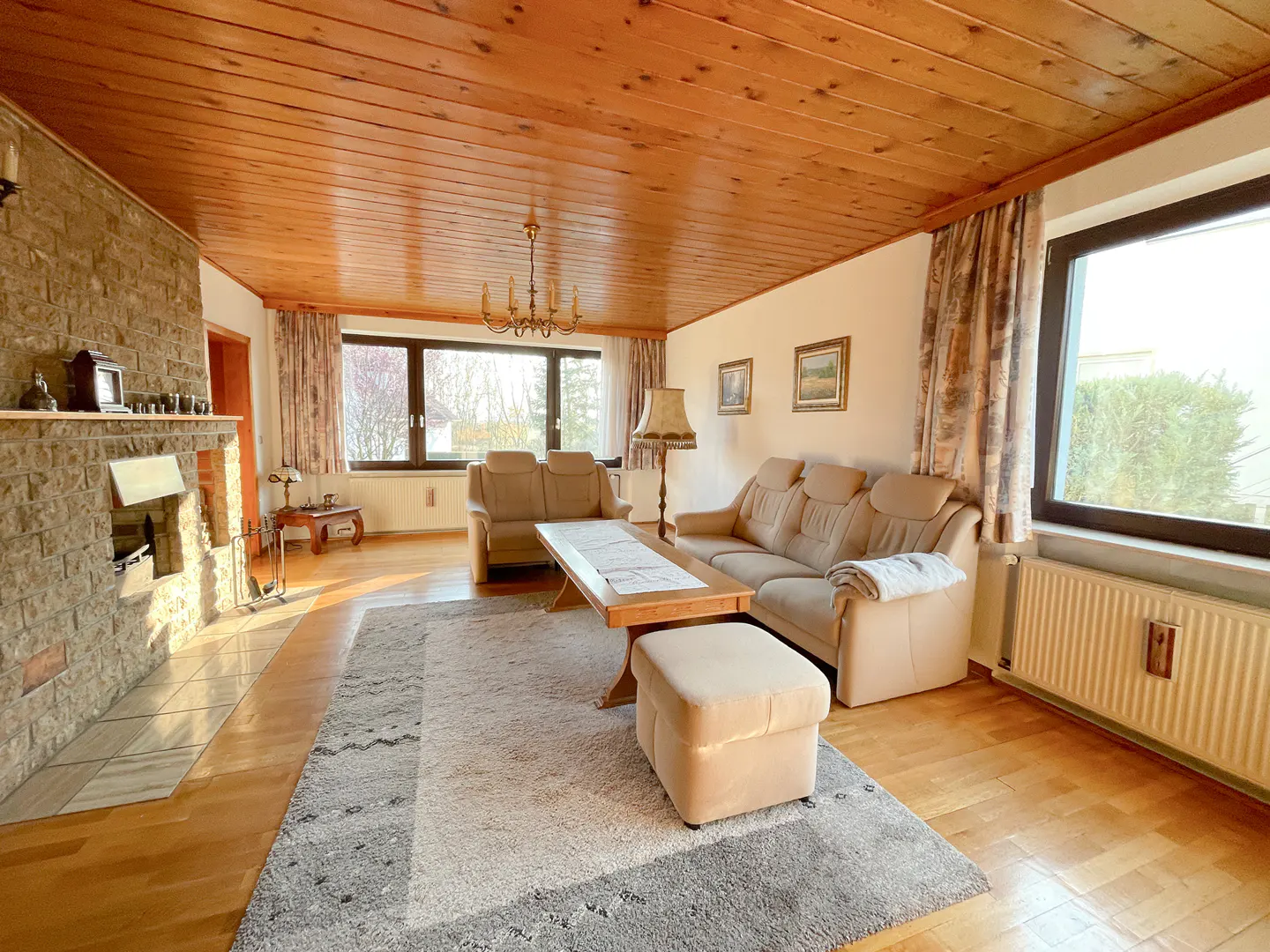 Living room with wood-paneled ceiling, stone fireplace, beige sofas, wood floors, and a gray rug. Windows offer natural light.