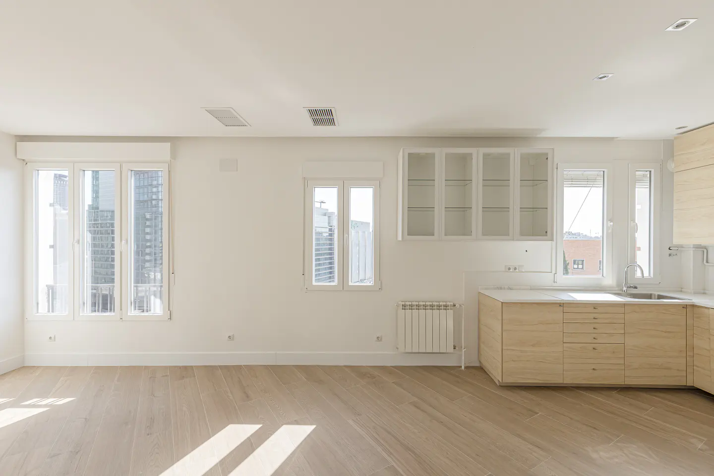 Bright, empty kitchen with light wood floors, white walls, and cabinets. Windows show city views.