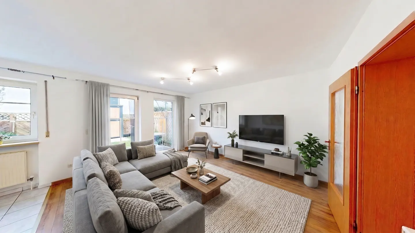 Bright living room with a gray sectional sofa, wood coffee table, TV, and a large rug. Windows overlook a green yard.