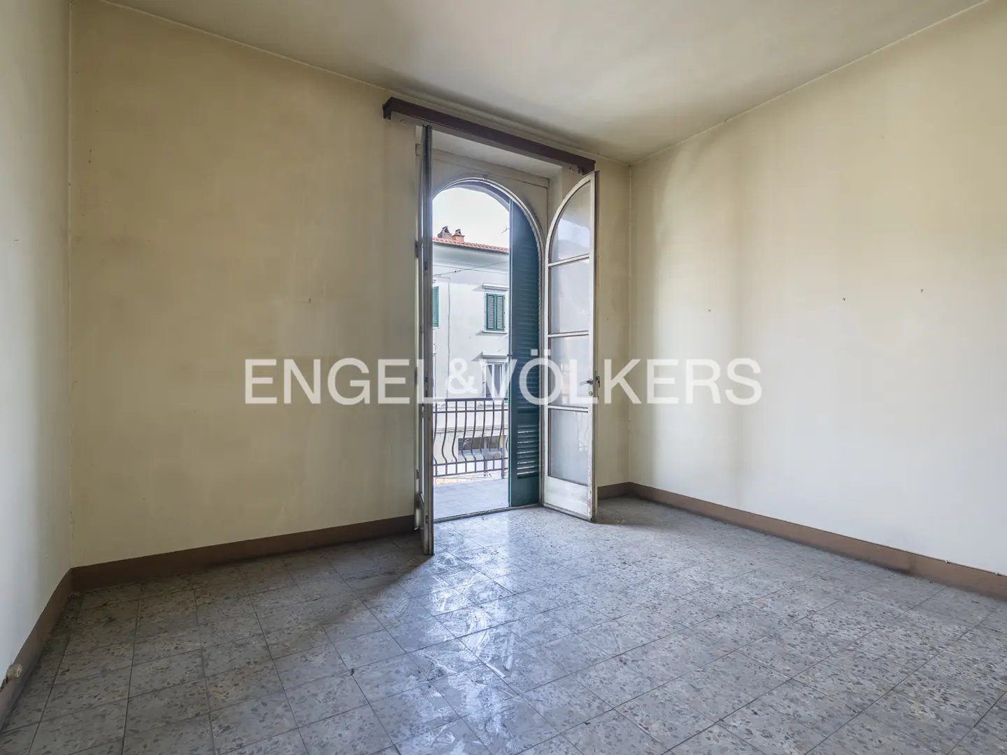 Empty room with beige walls and gray tiled floor. Open arched doorway leads to a balcony with a view of a building.