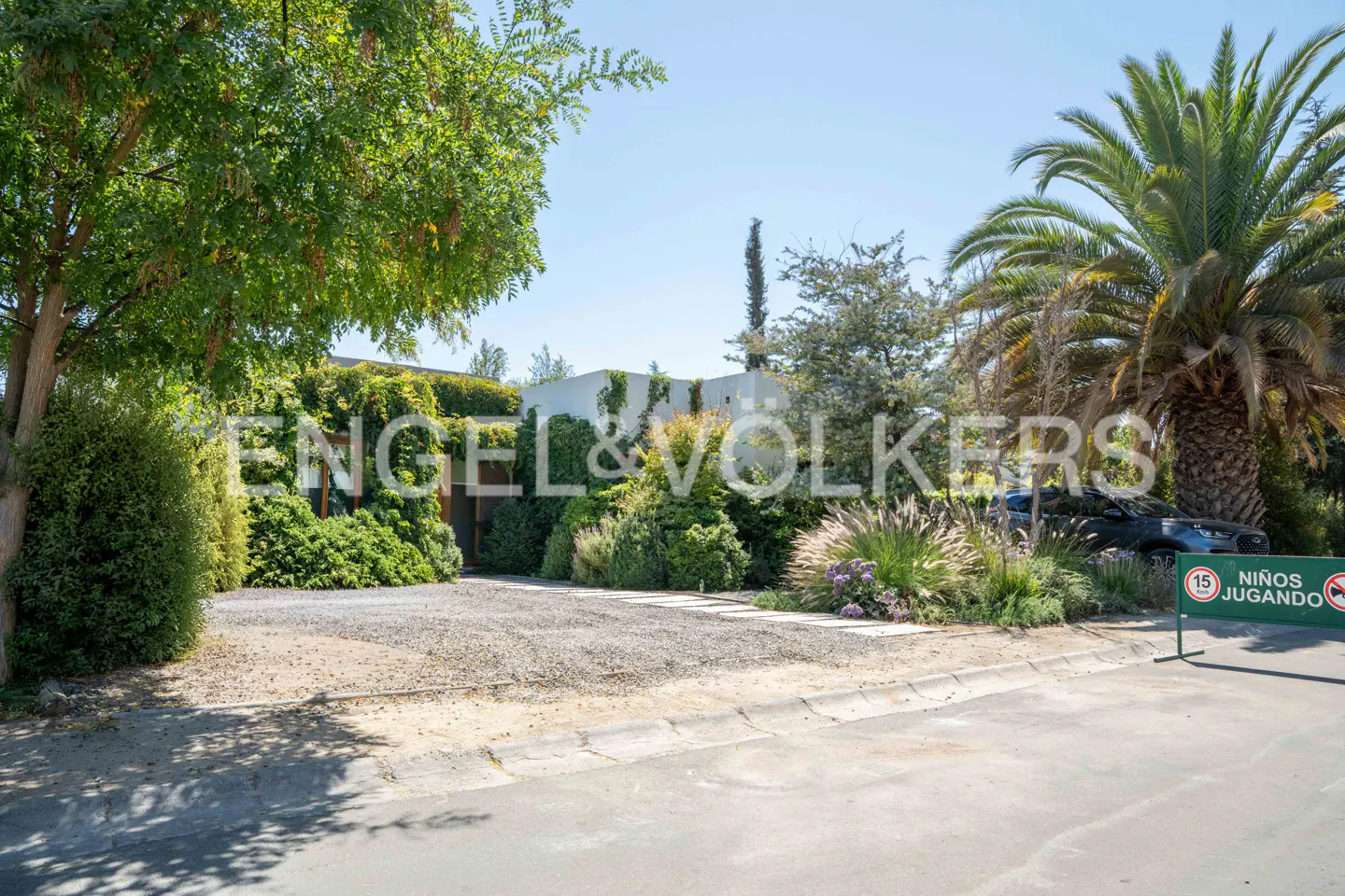 Exterior view of a modern white house with lush green landscaping and an Engel & Volkers sign. A gravel driveway leads to the house.