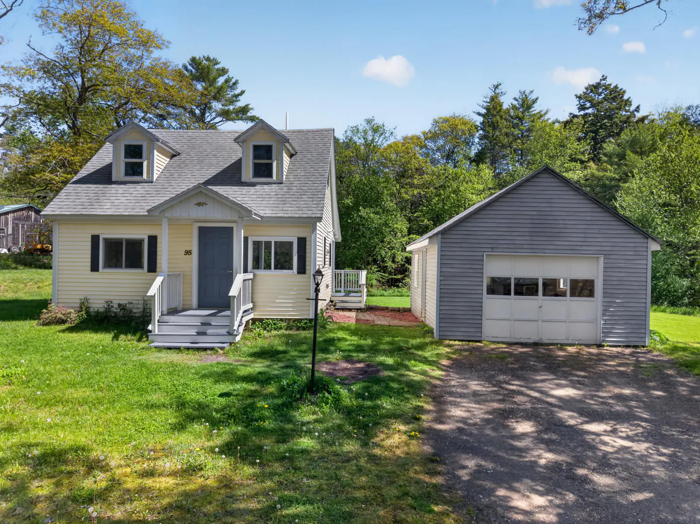 A yellow cottage-style house with a grey roof, dormer windows, and a detached grey garage set on a grassy, wooded lot.