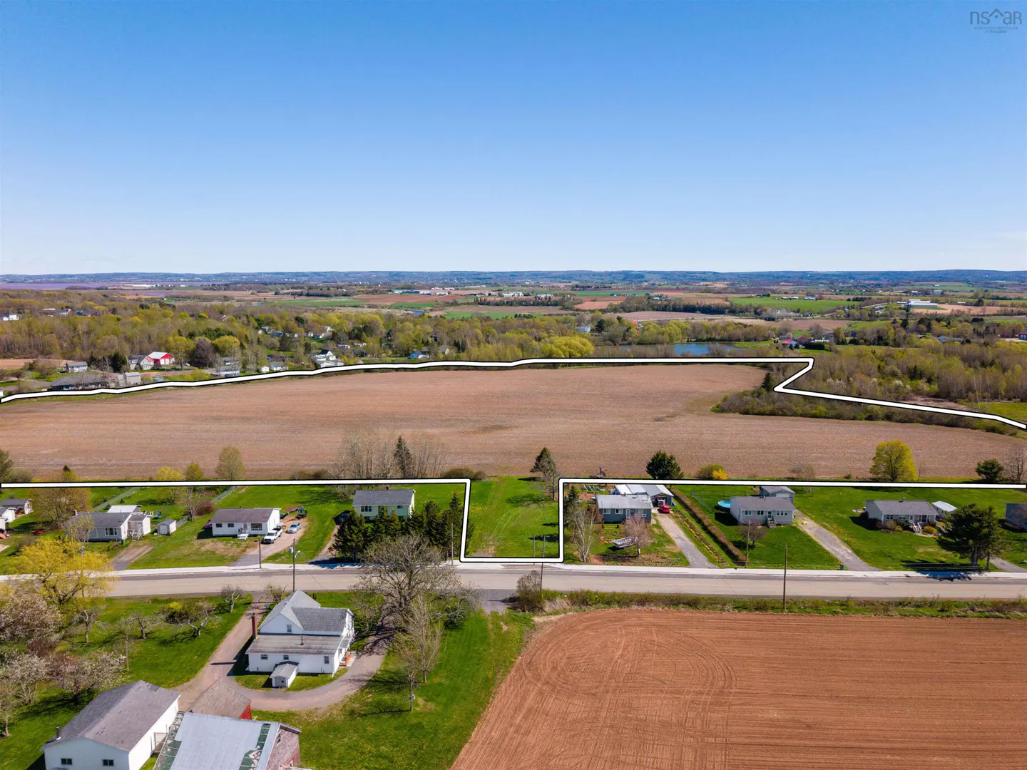 Aerial view of rural property with houses, green lawns, and brown fields under a clear blue sky.