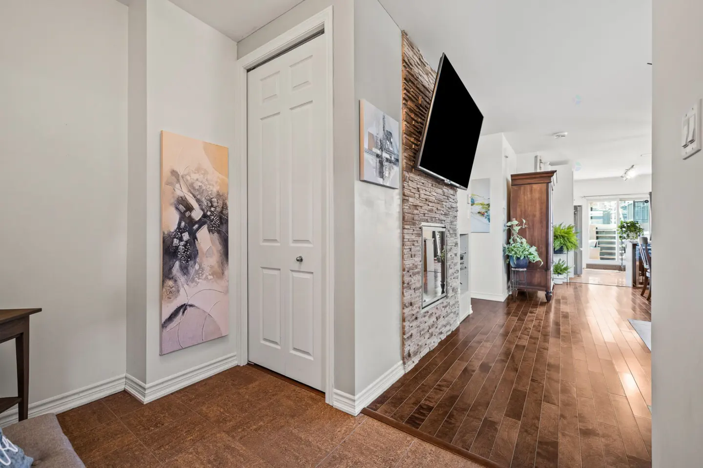 Hallway with tile floor, white door, abstract art, and a stone fireplace wall with a mounted TV leading to a living area.