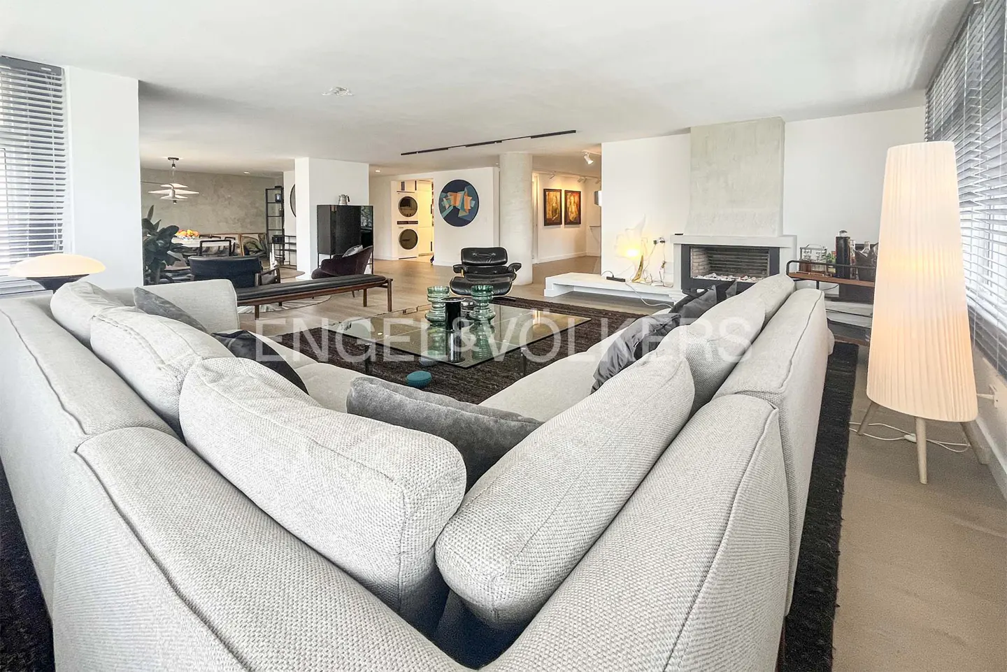 Wide shot of a modern living room with a large gray sectional sofa, glass coffee table, fireplace, and tall white lamp.